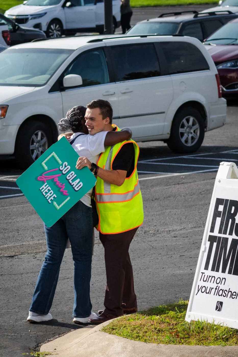 A parking lot greeter in a safety vest hugging a guest holding a So Glad You're Here sign